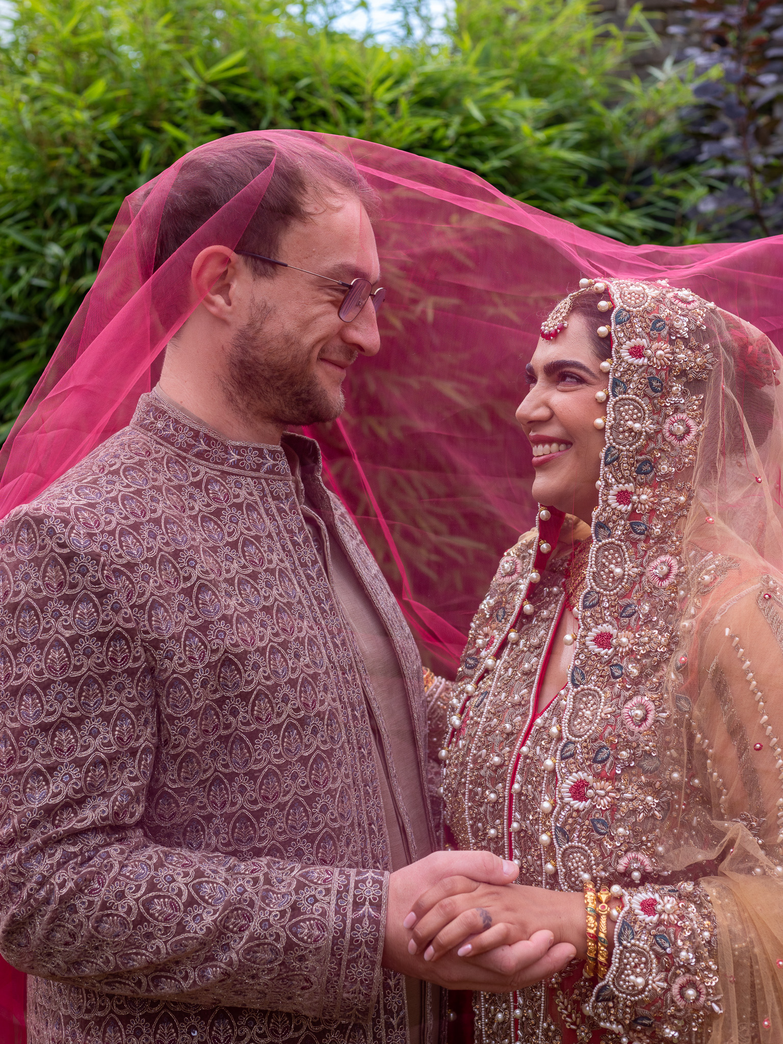 A smiling bride and groom stand close together under a pink veil, holding hands and gazing at each other. Both wear traditional embroidered wedding attire with intricate details, surrounded by greenery in the background.